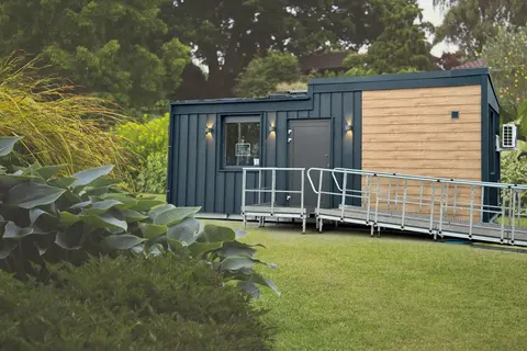 Außenansicht des TinyCareHome Modernes TinyCareHome-Modulgebäude mit barrierefreier Rampe in parkähnlicher Umgebung. Das kompakte Gebäude kombiniert dunkle Metallfassade mit Holzverkleidung und verfügt über eine Eingangstür mit Beleuchtung.