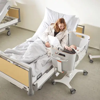 Mutter liegt im Krankenhausbett und kümmert sich um ihr Neugeborenes im höhenverstellbaren Säuglingsbett Jovie neben dem Bett in einem Patientenzimmer der Entbindungsstation.
