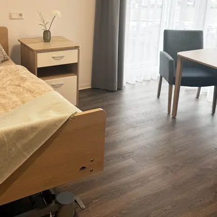 Habitación de residente con decoración moderna, con una acogedora cama de cuidados de Stiegelmeyer, mesita de noche y zona de descanso en una residencia de adultos mayores de Compassio
