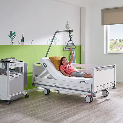 Dziecko leży w łóżku szpitalnym dla młodzieży Seta pro – wersja junior i korzysta z uchwytu trójkątnego na podnośniku