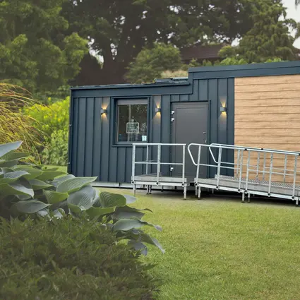 Modern TinyCareHome modular building with barrier-free ramp in park-like surroundings. The compact building combines a dark metal façade with wood panelling and has an entrance door with lighting.