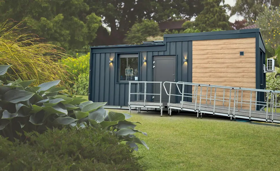 Modern TinyCareHome modular building with barrier-free ramp in park-like surroundings. The compact building combines a dark metal façade with wood panelling and has an entrance door with lighting.