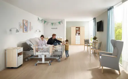 Familienzimmer auf einer Entbindungsstation mit einem wohnlichen Partnerbett und einem Babybett; Ein junges Paar sitzt im Bett und ein kleiner Junge steht vor dem Bett und spielt mit einerm Kuscheltier. Im Babybett liegt ein Neugeborenes und wird liebevoll von seiner Mutter angeschaut. Das Zimmer ist hell und gemütlich eingerichtet, mit Wickelkommode, Schrank, Tisch und Stühlen.