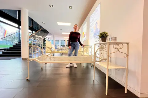 Historic beds and bedside cabinets in the Stiegelmeyer showroom in Herford. Amelie Plaßmann, a marketing communications apprentice at Stiegelmeyer, stands between the historic beds and bedside cabinets and smiles for the camera.
