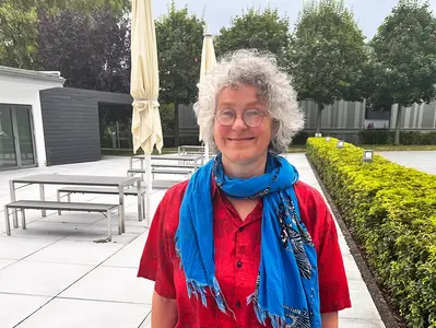 La Dra. Karen von Hünerbein posa sonriente en el patio del edificio administrativo de Stiegelmeyer en Herford. Lleva una blusa roja y un pañuelo azul.