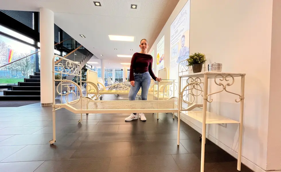 Historyczne łóżka i szafki nocne na wystawie Stiegelmeyer w Herford. Amelie Plaßmann, stażystka ds. komunikacji marketingowej w firmie Stiegelmeyer, stoi między historycznymi łóżkami i szafkami nocnymi i uśmiecha się do aparatu.