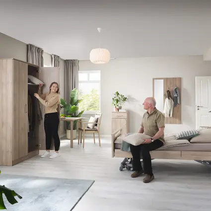 Luminosa y acogedora habitación de residente en una residencia de mayores con una cama asistencial, muebles de madera y grandes ventanas; una mujer abre un armario mientras un hombre mayor está sentado en la cama.