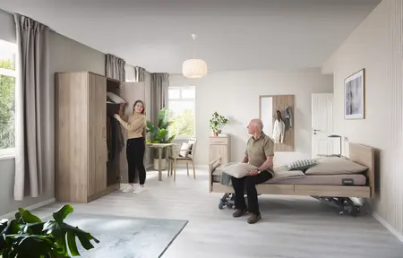 Luminosa y acogedora habitación de residente en una residencia de mayores con una cama asistencial, muebles de madera y grandes ventanas; una mujer abre un armario mientras un hombre mayor está sentado en la cama.