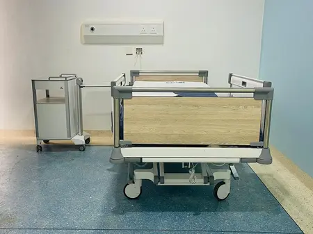 Łóżko szpitalne i szafka przyłóżkowa firmy Stiegelmeyer znajdują się w dużej niebieskiej sali dla pacjentów.