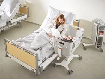 La madre está tumbada en la cama del hospital y cuida de su recién nacido en la cuna Jovie, de altura regulable, situada junto a la cama en una habitación de la sala de maternidad.
