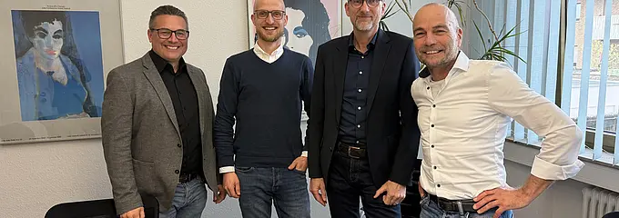 Four men are standing close together in a bright office or meeting room. They are smiling at the camera; in the foreground, a contract folder can be seen on a table.