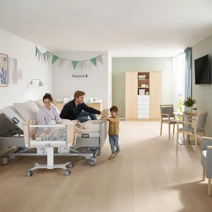 Habitación familiar en una maternidad con una cómoda cama para la pareja y una cuna; una joven pareja está sentada en la cama y un niño pequeño está de pie delante de la cama jugando con un peluche. En la cuna hay un recién nacido al que su madre mira con cariño. La habitación es luminosa y está amueblada de forma acogedora, con cambiador, armario, mesa y sillas.