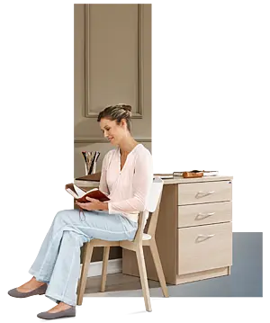 Comprehensive furniture solutions from Stiegelmeyer A blonde woman is sitting on a wooden chair at a small desk, reading a notebook; on the table are pens in a cup and other writing materials, in a bright, homelike environment.